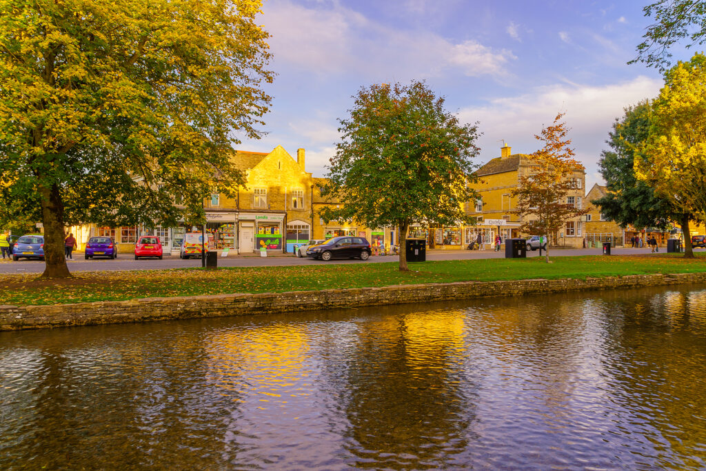 Bourton-on-the-Water, UK - October 17, 2022: Sunset scene of typical houses, the river Windrush, locals and visitors, in the village Bourton-on-the-Water, the Cotswolds region, England, UK